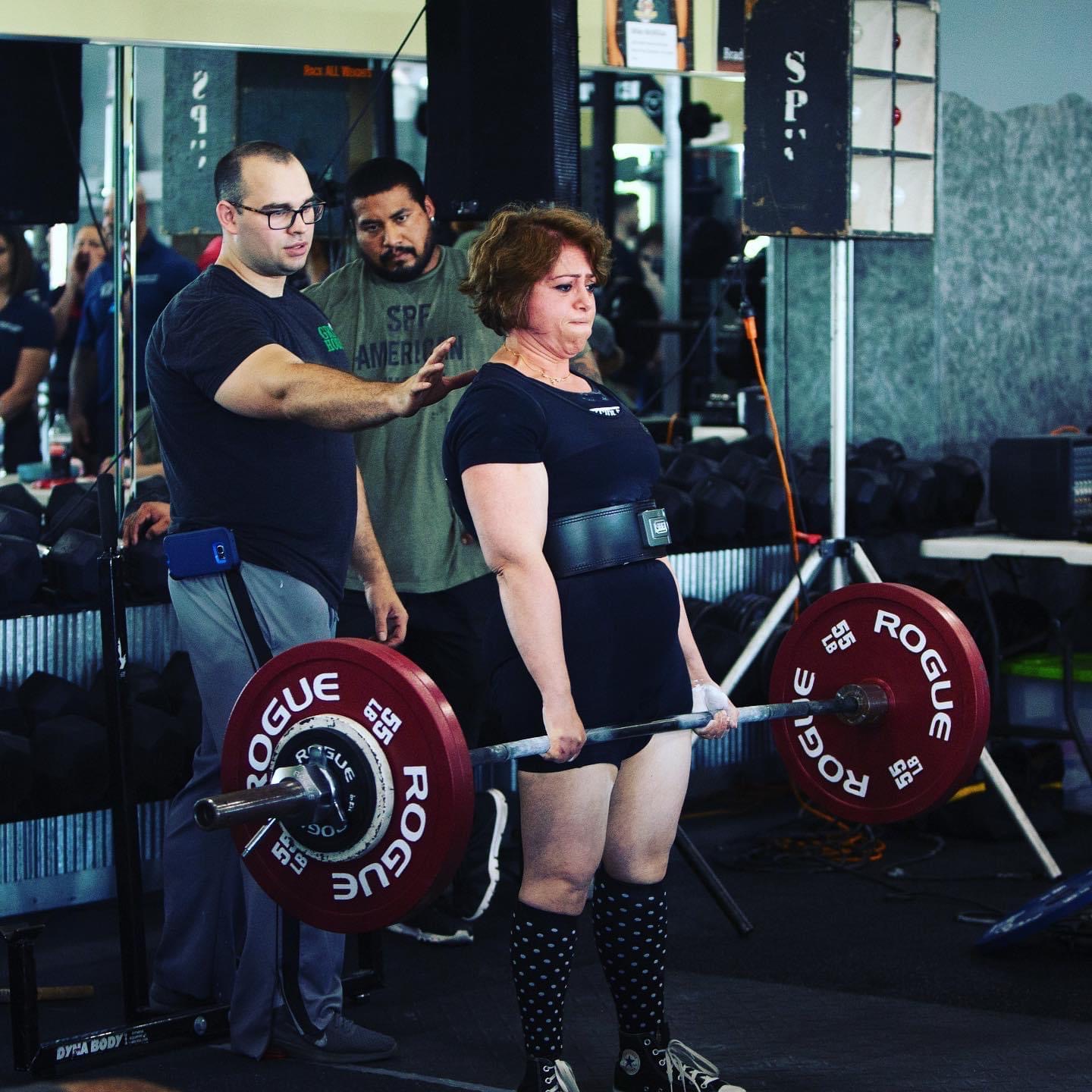 Champion powerlifter Anjuli Bhat-Mejia performs a move at a CrossFit competition while a spotter assists.